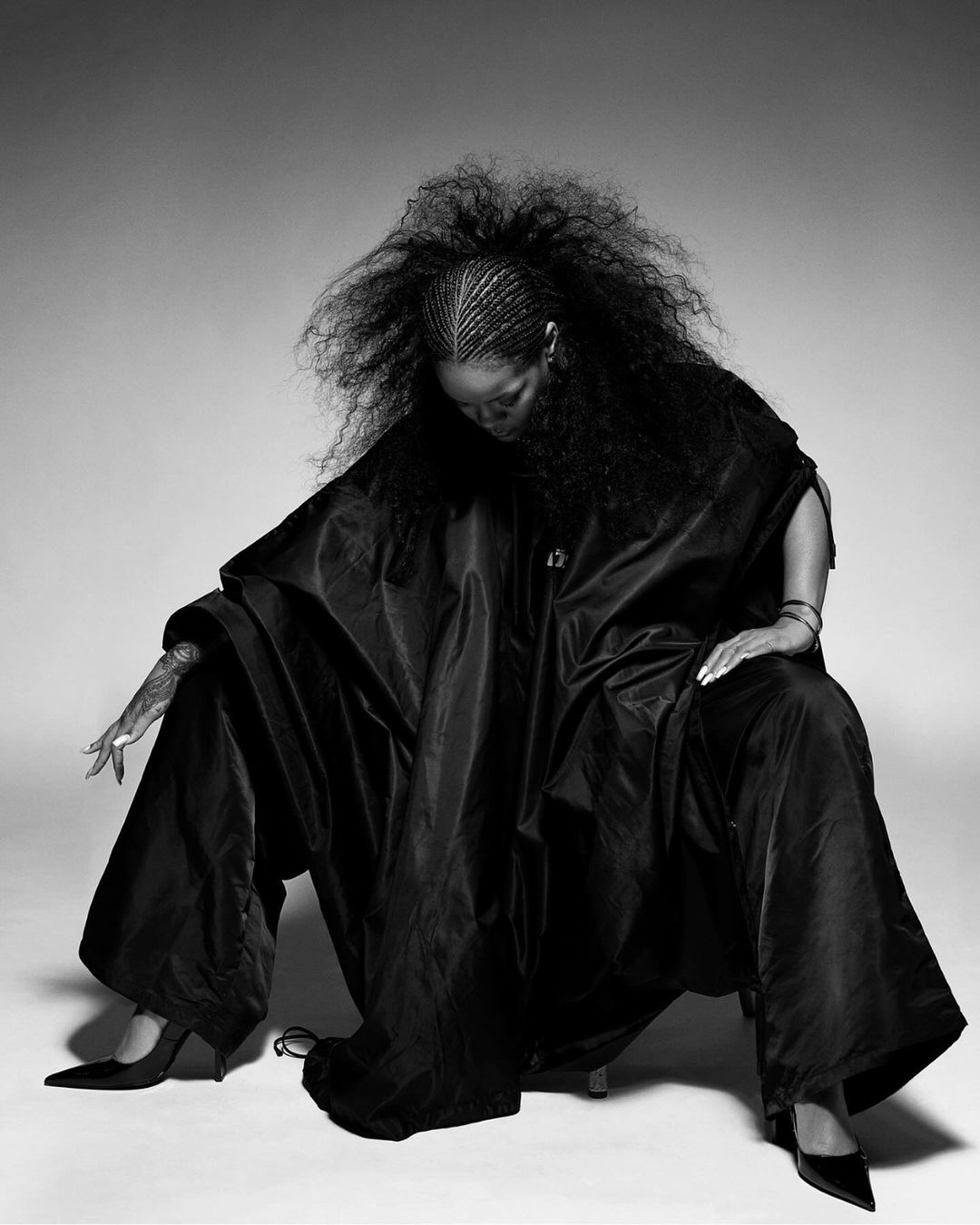 Black and white studio portrait of an adult woman with braids and afro-textured hair wearing a large black satin poncho and heels.
