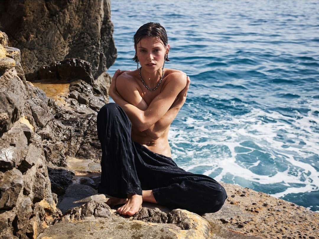 A woman sits on rocks beside the sea, hugging herself, while gazing directly at the camera in a beauty or fashion shot.