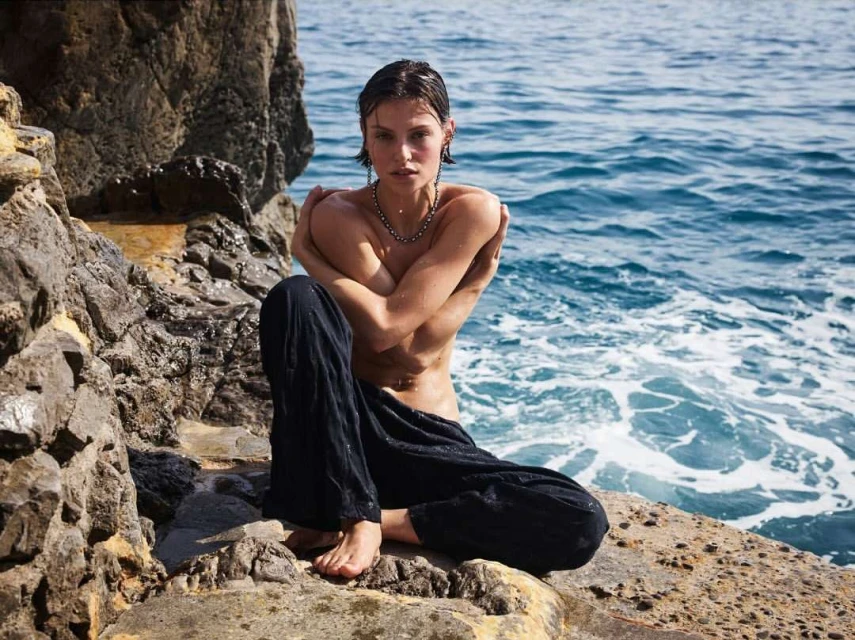 A woman sits on rocks beside the sea, hugging herself, while gazing directly at the camera in a beauty or fashion shot.