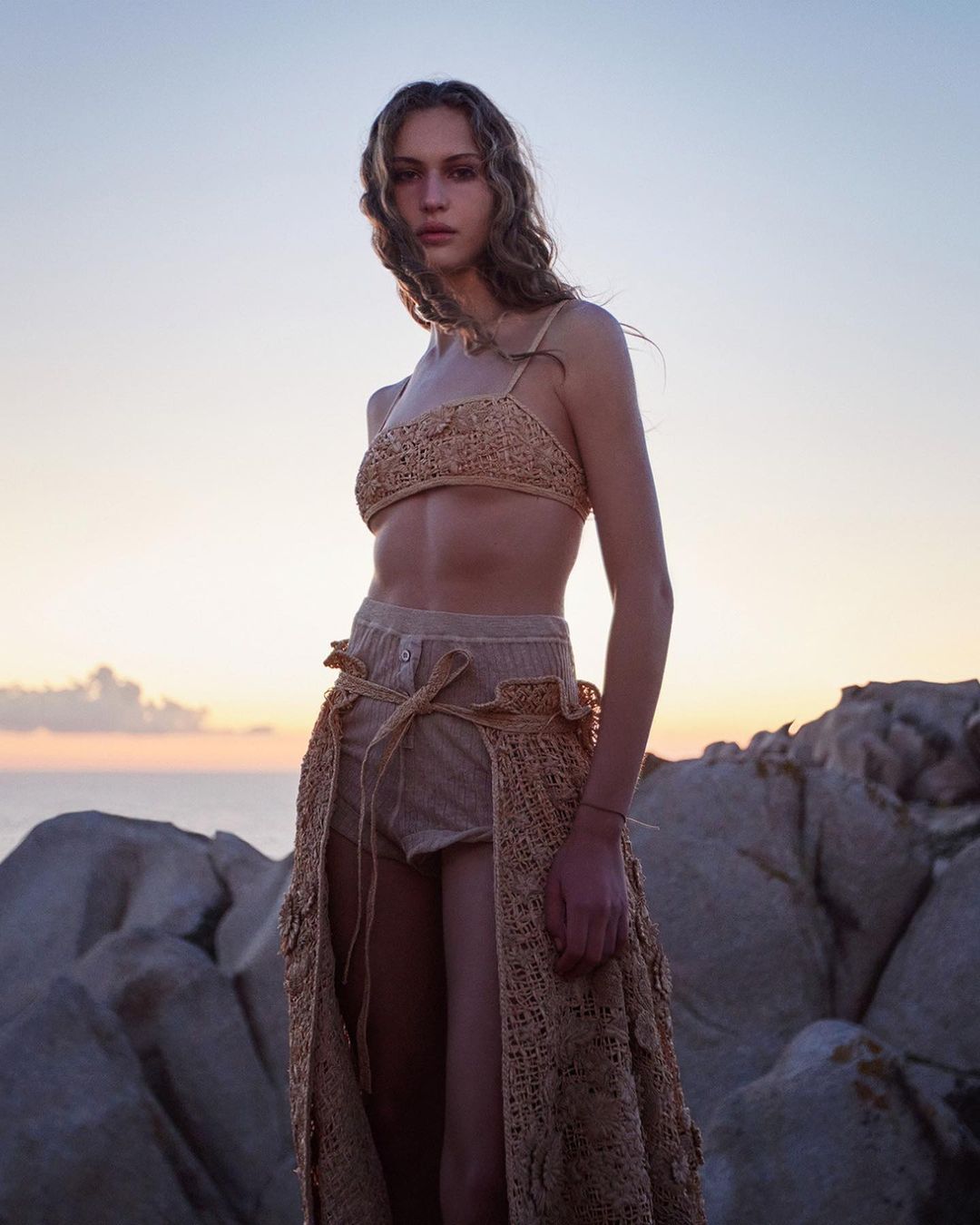 A woman poses wearing a tan crocheted bikini top and skirt, standing on rocks by the sea with a colorful sky at dusk.