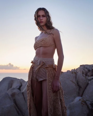 A woman poses wearing a tan crocheted bikini top and skirt, standing on rocks by the sea with a colorful sky at dusk.