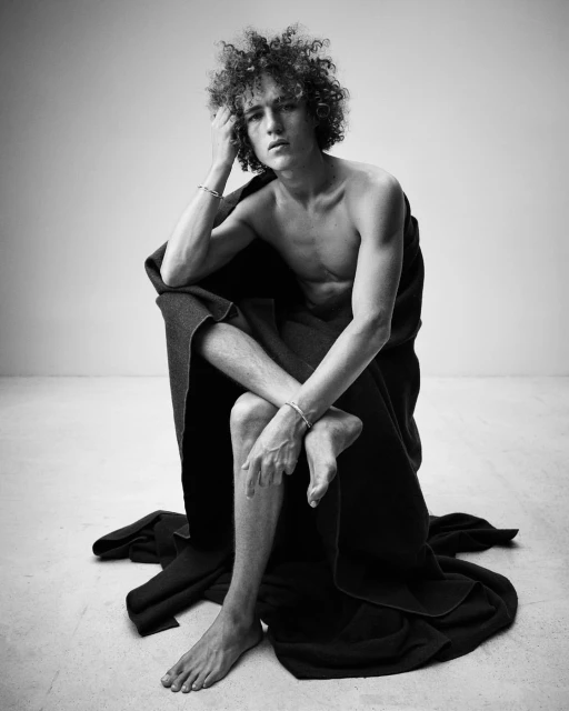 Black and white studio portrait of a young adult male with curly hair, wrapped in a dark blanket, seated with arms and legs crossed.