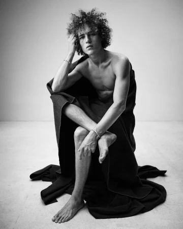 Black and white studio portrait of a young adult male with curly hair, wrapped in a dark blanket, seated with arms and legs crossed.