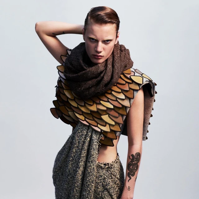 Stylish person with short hair poses in a distinctive woven garment with shoulder armor, a large scarf, and a snake tattoo.