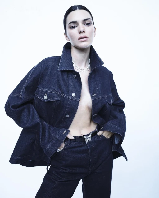Fashion portrait of a woman with dark hair wearing a denim jacket and jeans against a white background in a photo studio.