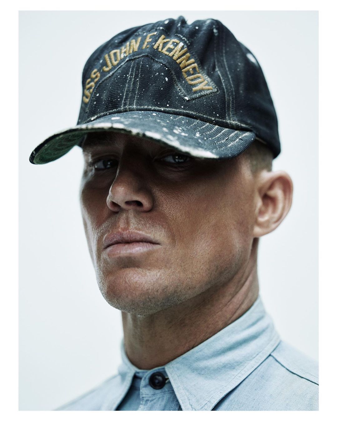 Close-up portrait of a man wearing a dark distressed baseball cap with gold lettering and a light blue buttoned shirt against a white wall.