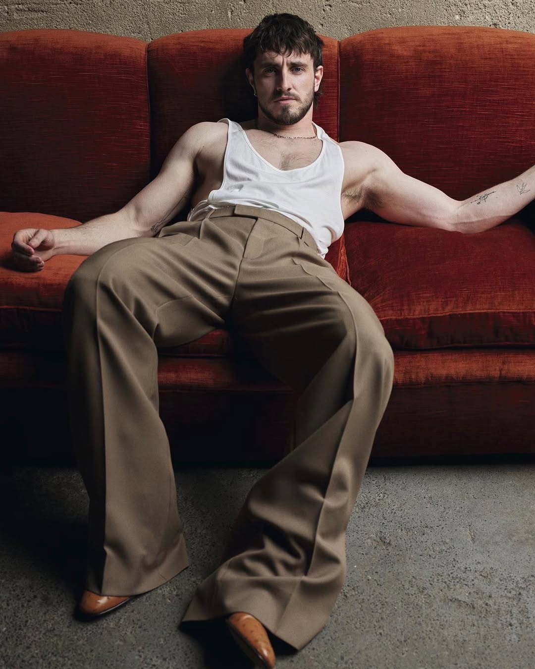 Paul Mescal lounges on a velvet couch wearing a white tank top and wide-legged khaki pants, showcasing a relaxed and fashionable pose.