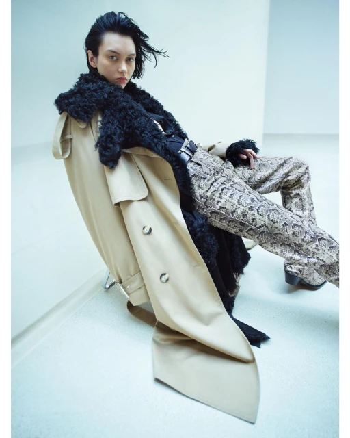 Editorial fashion shot of an androgynous model wearing a trench coat with fur collar and snakeskin pants in a studio setting.