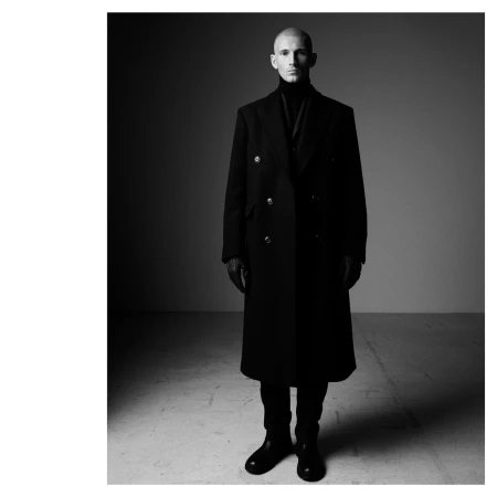 Bald male model in a long dark coat and turtleneck standing against a plain studio background