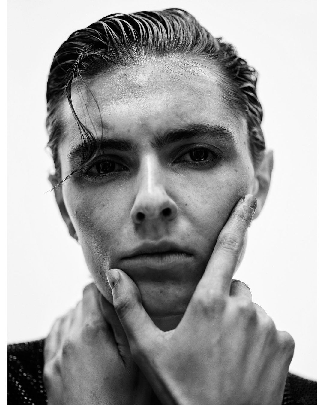 Close-up black and white portrait of a person with slicked-back dark hair holding hand to chin