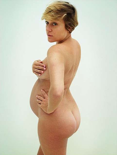 Nude pregnant woman with blonde hair poses in a studio, facing the viewer with a hand on her hip and another covering her breasts.