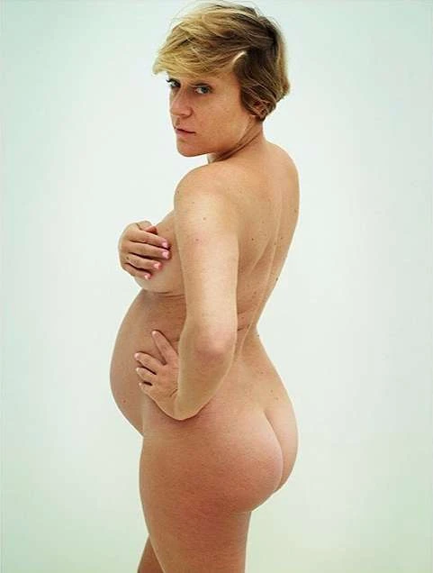 Nude pregnant woman with blonde hair poses in a studio, facing the viewer with a hand on her hip and another covering her breasts.