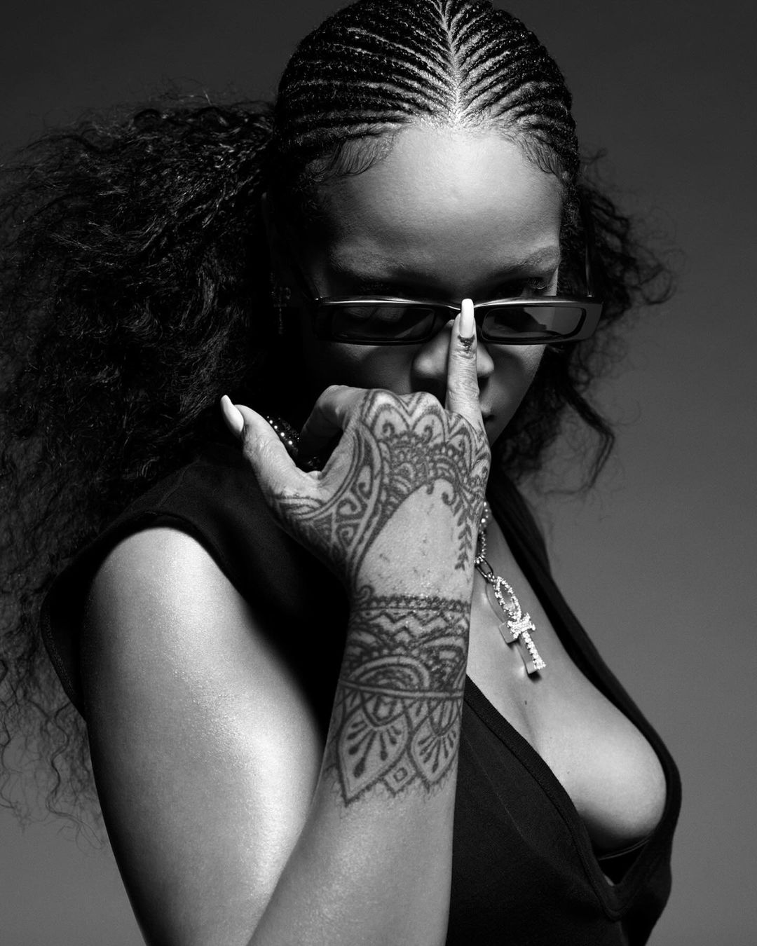 Black and white portrait of an adult female wearing sunglasses, sleeveless top, necklace, and a hand tattoo against a solid background.