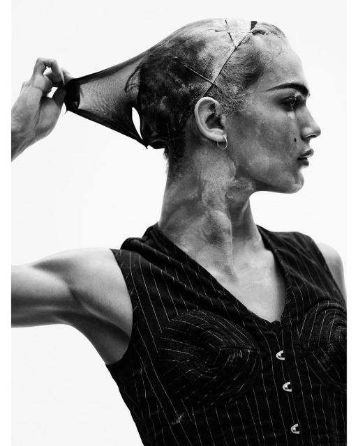 Monochrome portrait of adult with mesh fabric over head, showcasing textured skin, earring, and pinstripe vest against light backdrop.