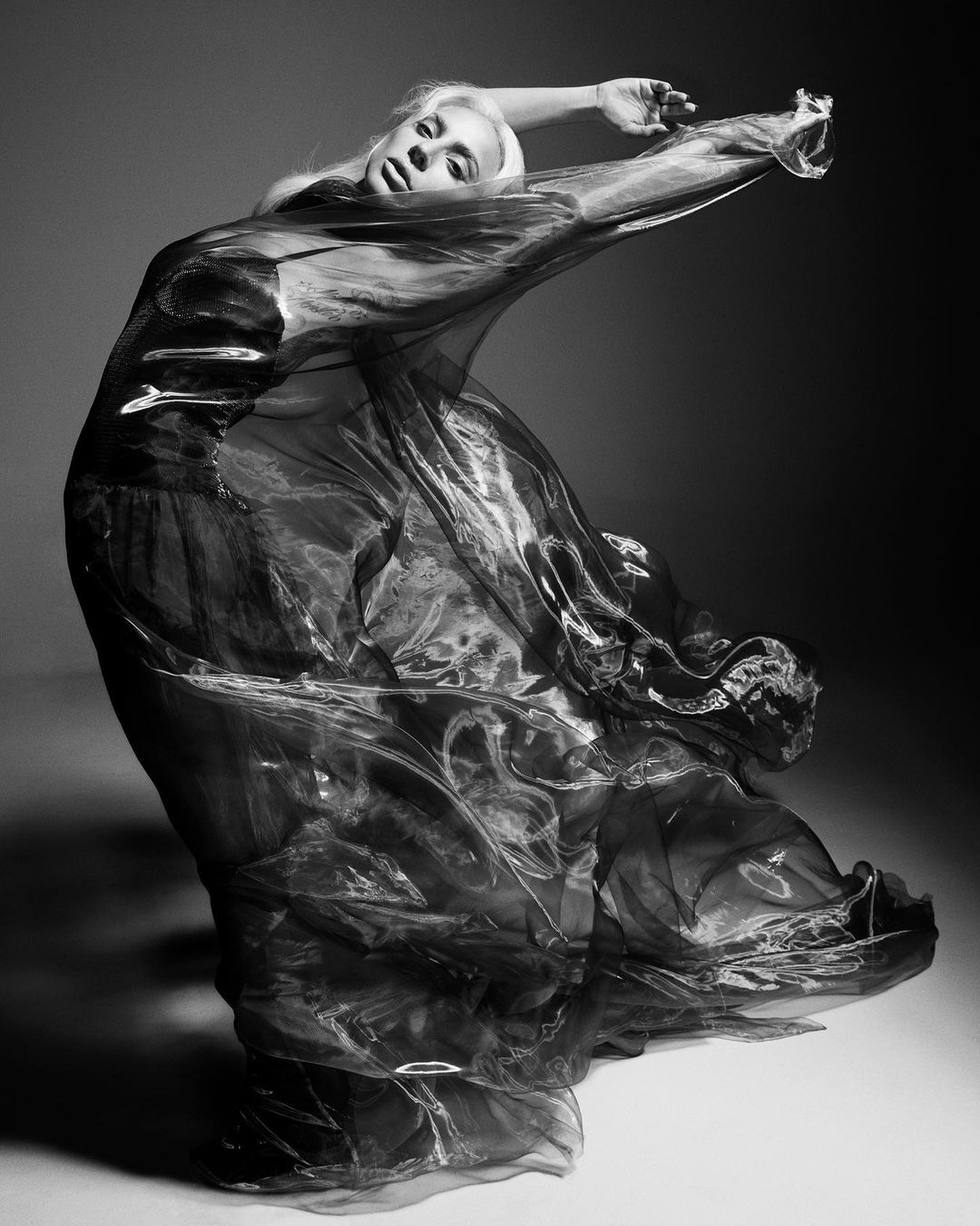 Monochrome portrait of a female model in flowing fabric, striking a dynamic pose in a studio setting with soft, diffused lighting.