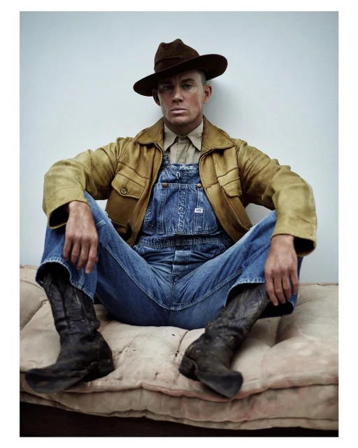Portrait of a man in overalls, brown jacket, and cowboy boots sitting on a tan cushion against a light gray studio backdrop, eye-level shot.