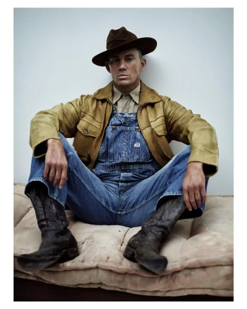 Portrait of a man in overalls, brown jacket, and cowboy boots sitting on a tan cushion against a light gray studio backdrop, eye-level shot.