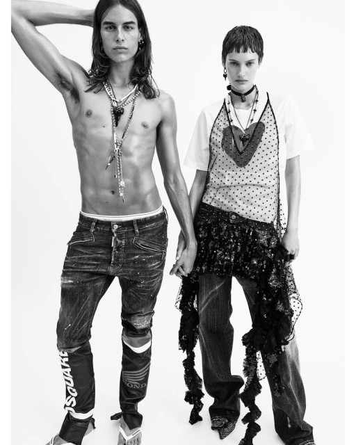 Black-and-white fashion shot of a shirtless man with long hair and a woman with short hair holding hands in front of a white background.