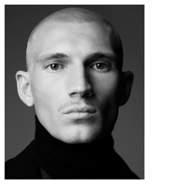 Black and white portrait of a young adult male with a shaved head, wearing a black turtleneck sweater, posing indoors.