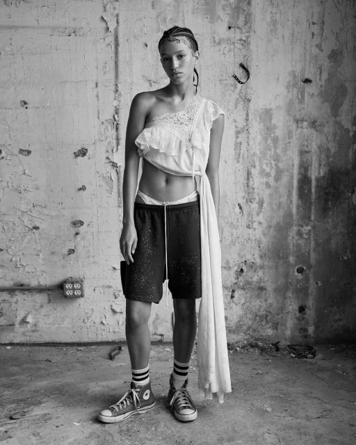 An adult female poses in an abandoned building, wearing a white top, shorts, socks, and sneakers, against a concrete wall with peeling paint.