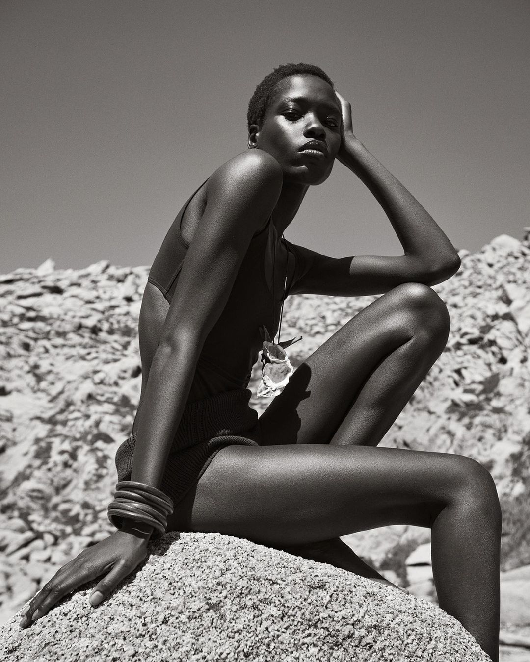 Black and white portrait of a Black woman sitting on a rock pile, posing in a swimsuit, with an open sky in the background.