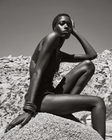Black and white portrait of a Black woman sitting on a rock pile, posing in a swimsuit, with an open sky in the background.