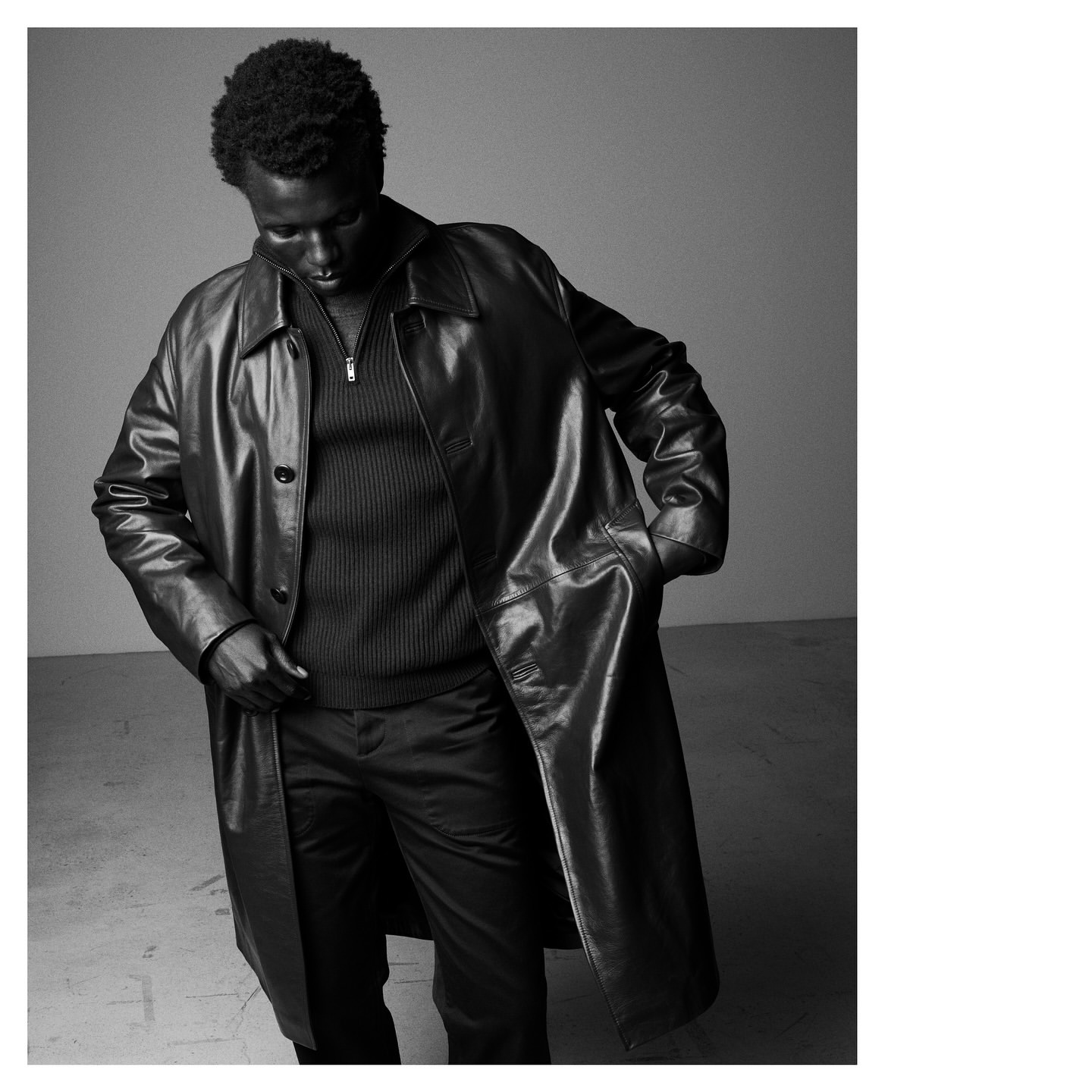 Black and white portrait of an African American male fashion model in a long leather trench coat, indoors, hands in pockets.