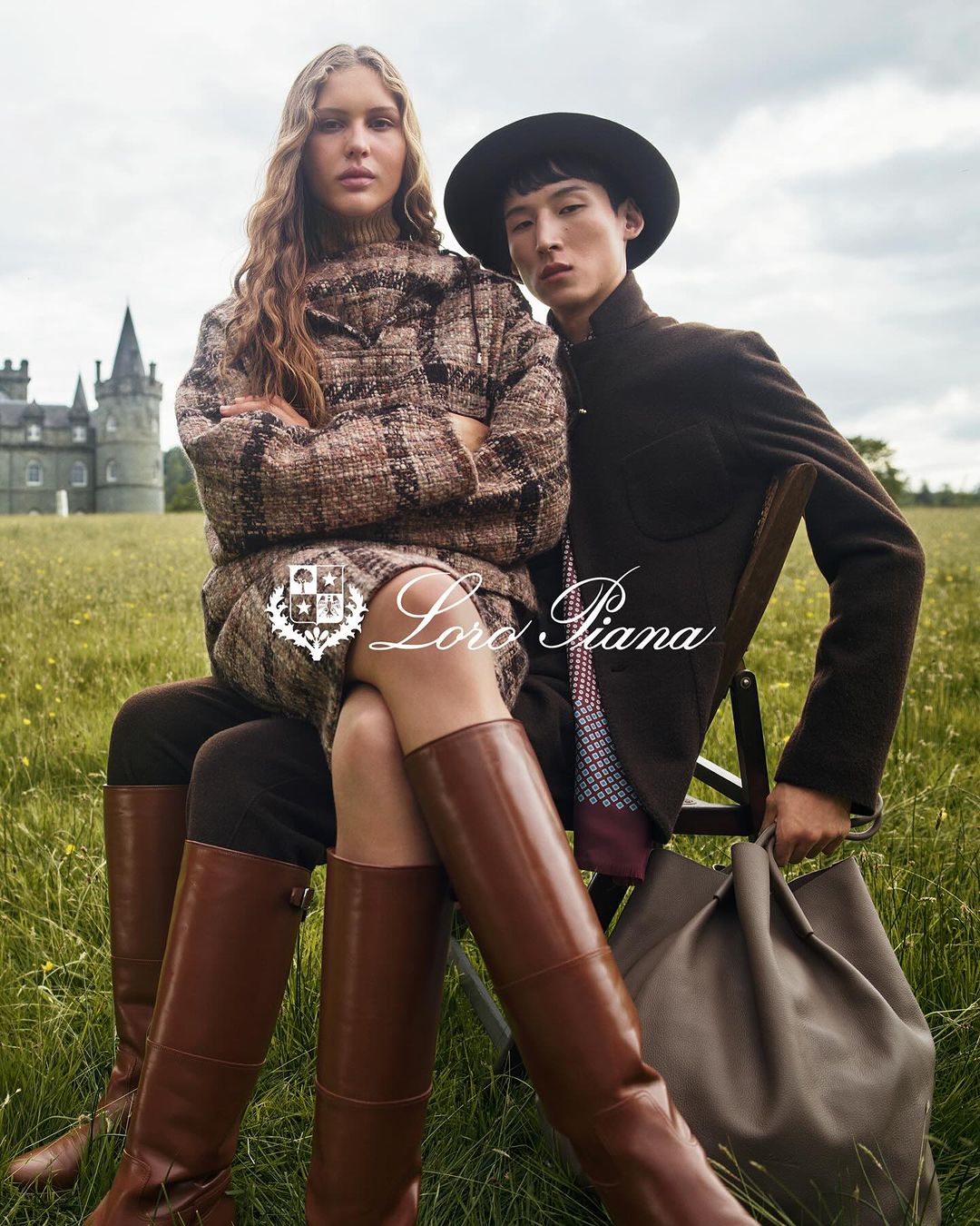Two models, an adult woman and an adult man, are posed outdoors in riding boots, skirts, jackets with a castle in the background.
