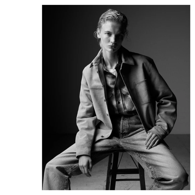 Fashion portrait of a woman with blonde hair, wearing a jacket and jeans, posing on a stool in a studio with dark background.