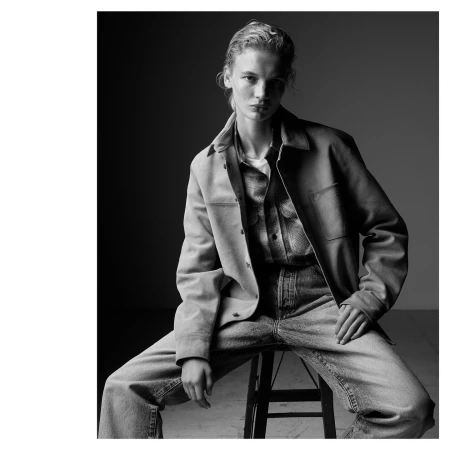 Fashion portrait of a woman with blonde hair, wearing a jacket and jeans, posing on a stool in a studio with dark background.