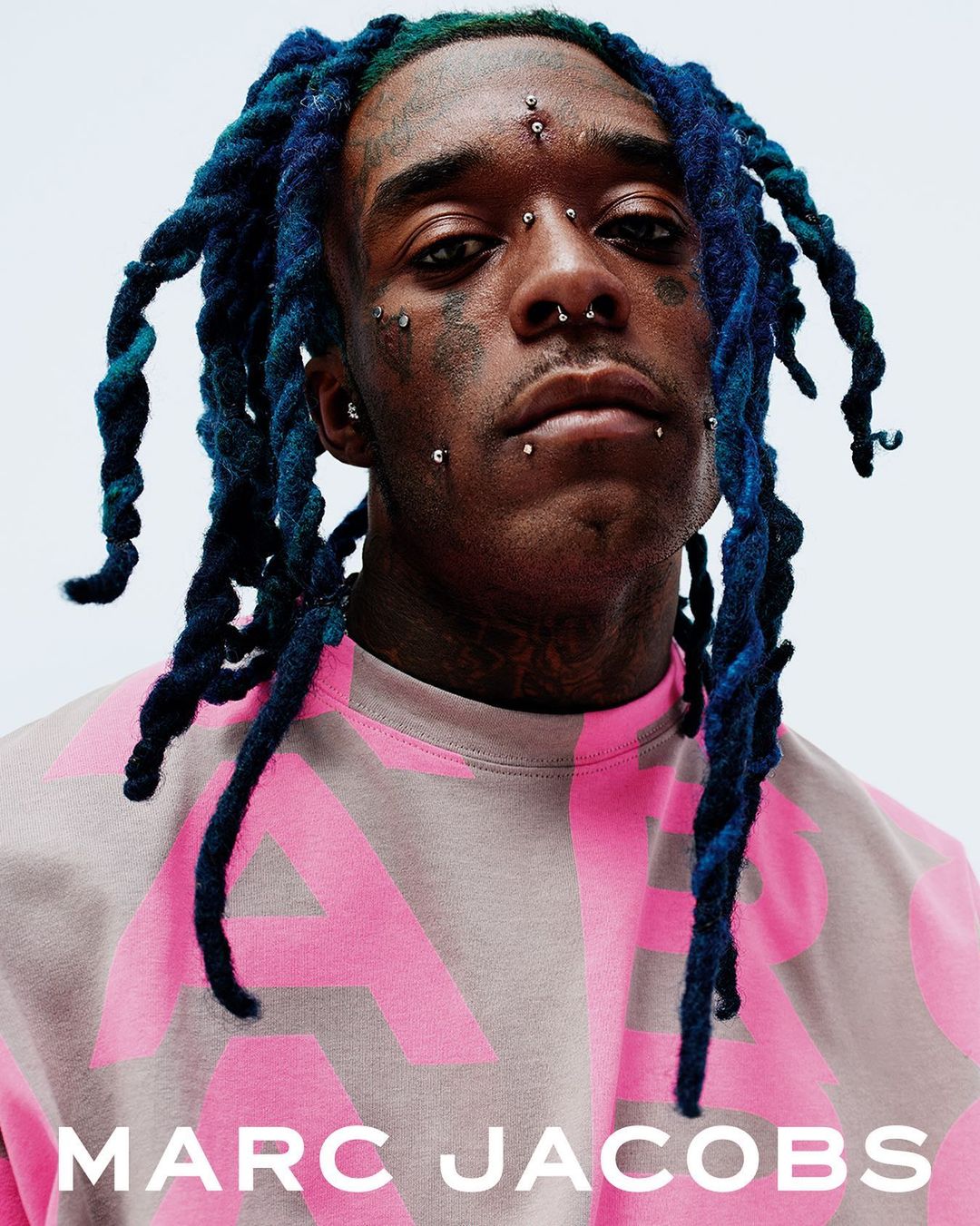 Close-up portrait of an adult male wearing a pink Marc Jacobs sweatshirt, featuring blue dreadlocks and multiple facial piercings.