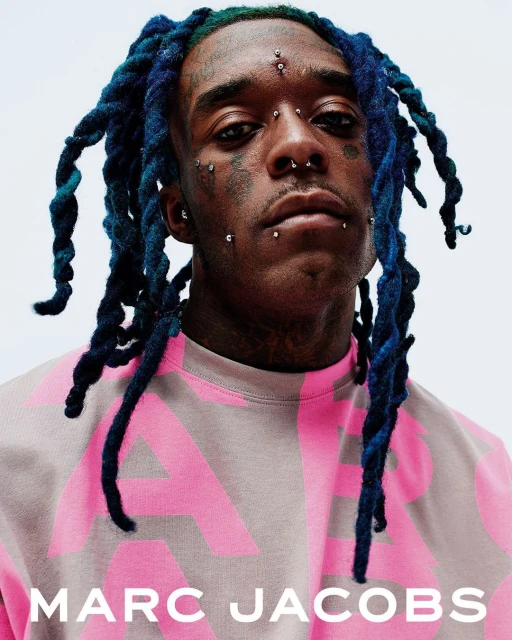 Close-up portrait of an adult male wearing a pink Marc Jacobs sweatshirt, featuring blue dreadlocks and multiple facial piercings.