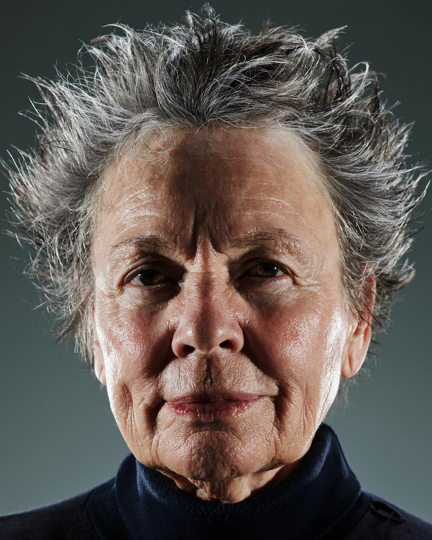 Close-up portrait of an elderly adult female with grey hair, wearing a dark turtleneck shirt against a dark grey studio backdrop.