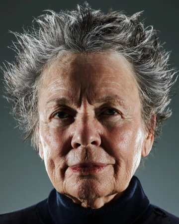 Close-up portrait of an elderly adult female with grey hair, wearing a dark turtleneck shirt against a dark grey studio backdrop.