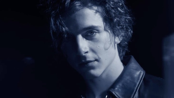 Close-up of a young adult male with curly dark hair and a leather jacket posing in a low-lit indoor setting with a serious expression.