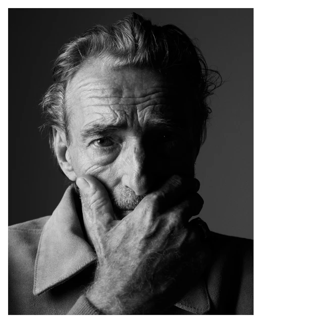 Black and white portrait of an elderly man with hand on his face, showing wrinkles and thoughtful expression, set against a grey backdrop.