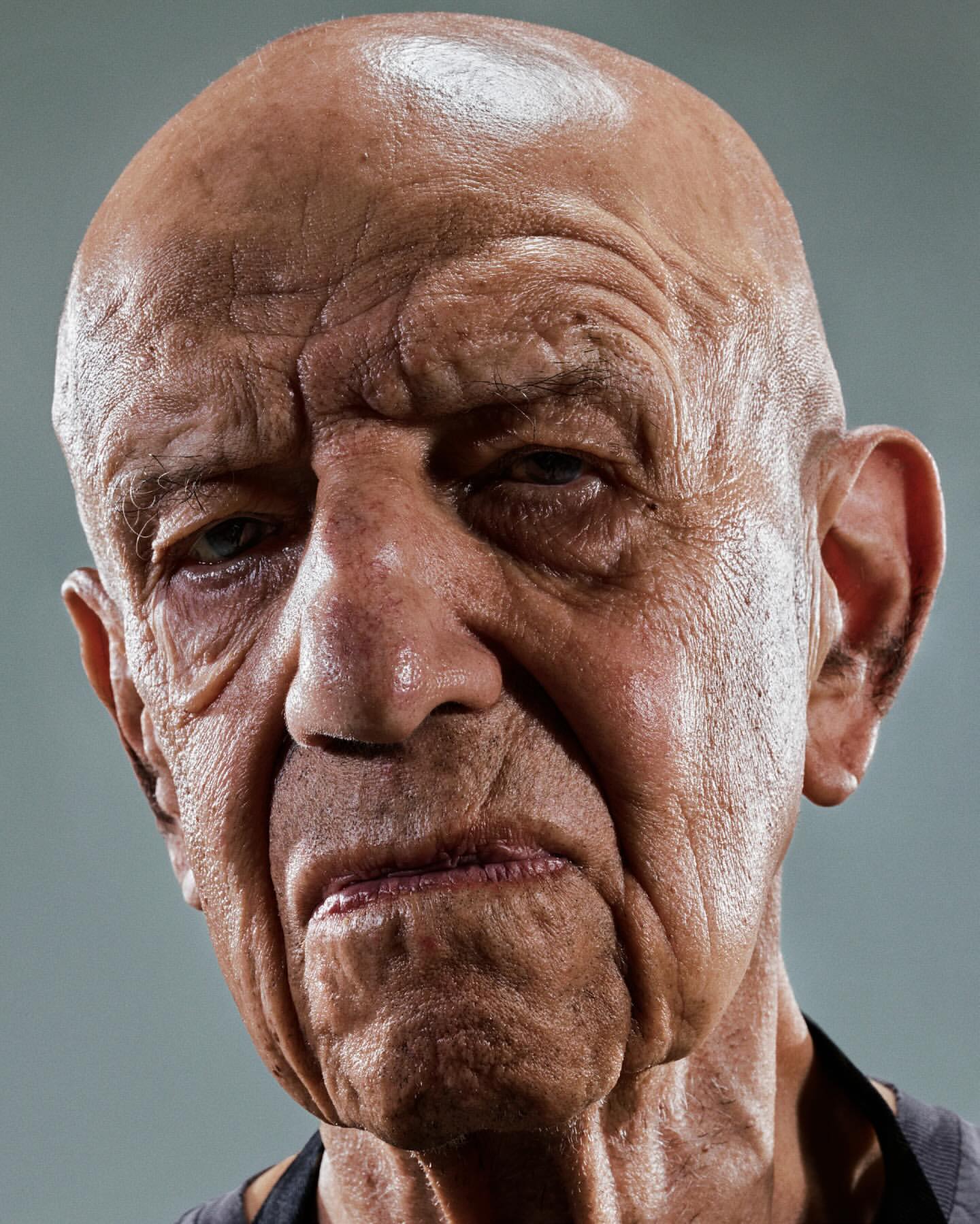 Close-up studio shot of a senior adult male with a shaved head and visible wrinkles, captured with soft lighting on a grey background.