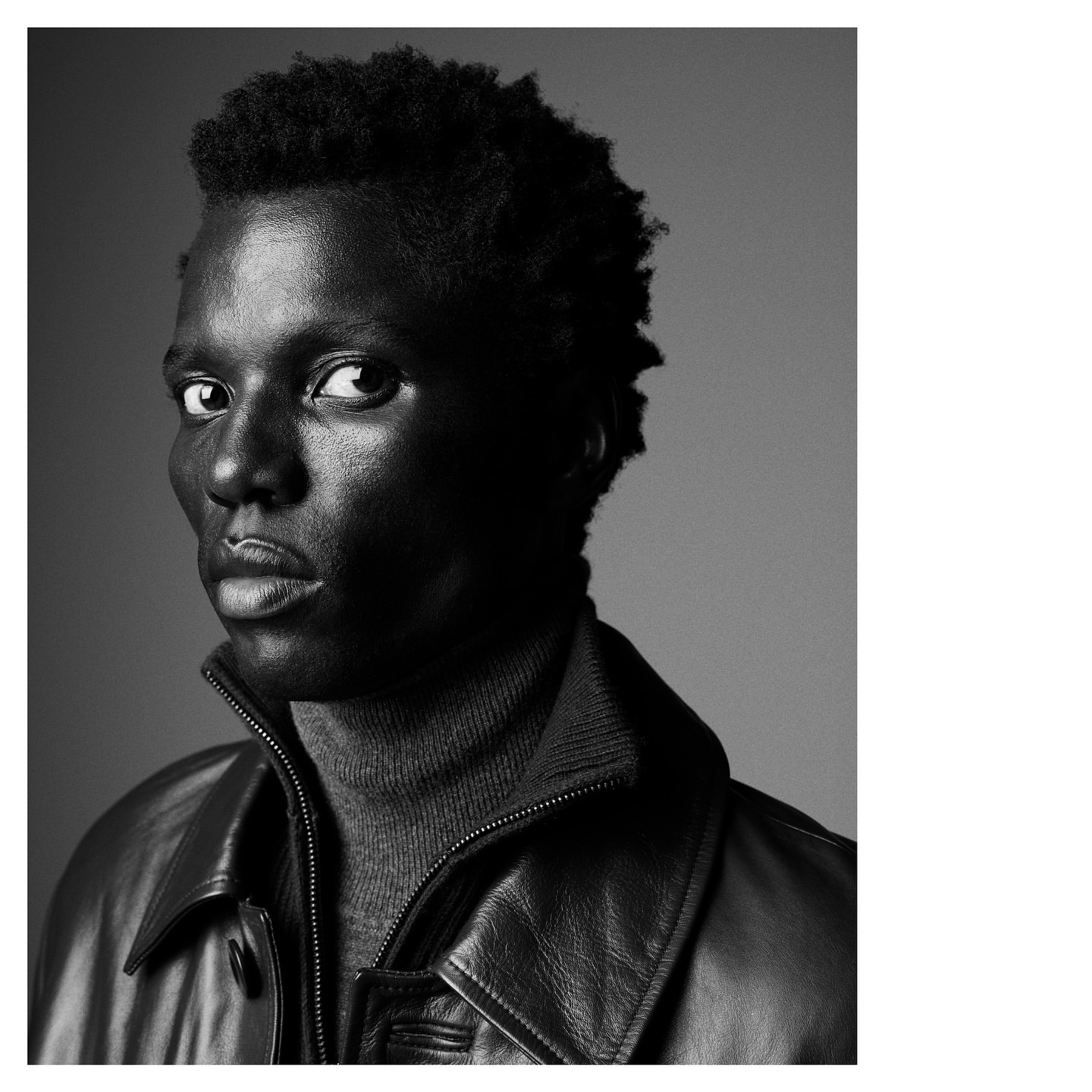 Portrait of a Black male model wearing a grey turtleneck and black leather jacket in a photo studio against a grey backdrop.