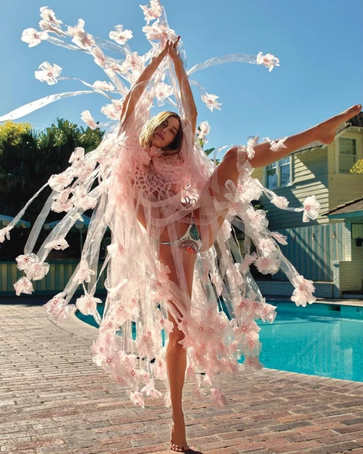 Fashionable woman in floral gown strikes a pose near the poolside of a house, basking in natural light under a clear blue sky.