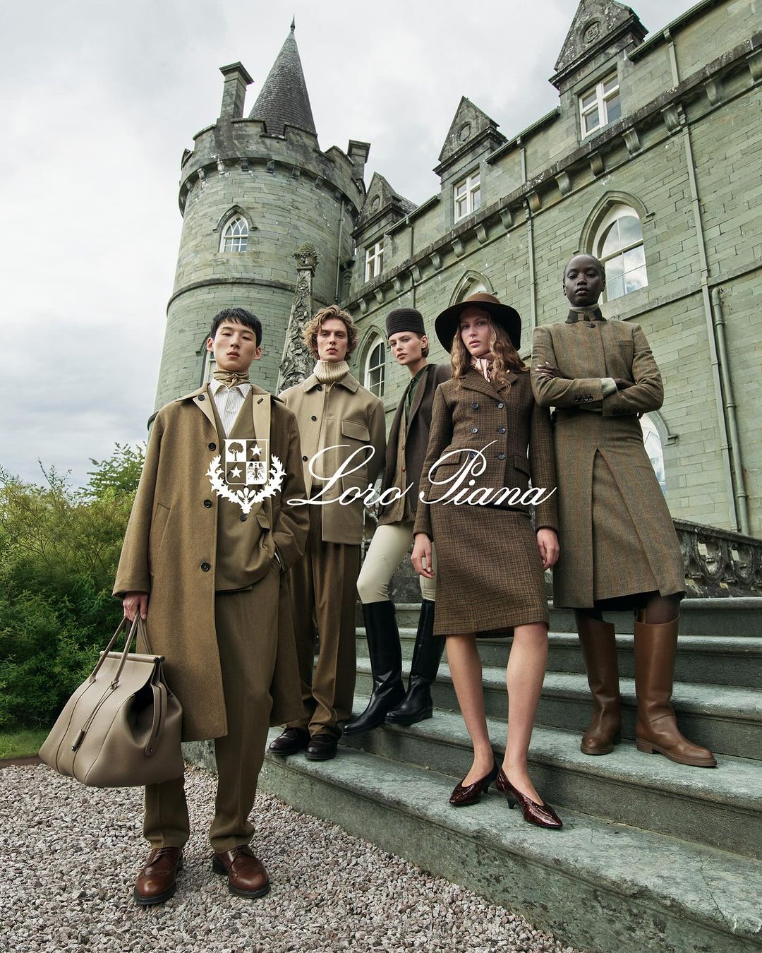 A group of five fashion models on stone steps in front of a historic castle, showcasing Loro Piana clothing against a green, outdoor backdrop.