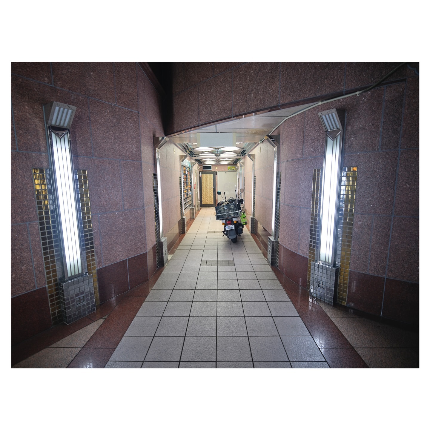 An indoor hallway with red and brown granite walls, tile flooring, fluorescent lighting, and a parked scooter at the far end.