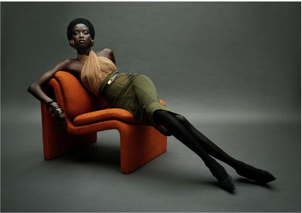 Adult woman with dark skin wearing a tan top and olive green skirt sitting on an orange chair in a photo studio with grey background.