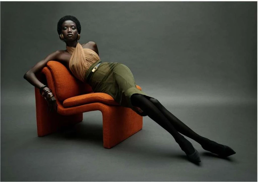 Adult woman with dark skin wearing a tan top and olive green skirt sitting on an orange chair in a photo studio with grey background.