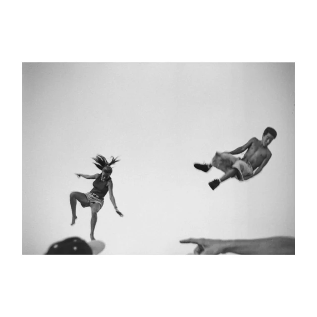 Monochromatic shot of two people mid-air, captured from a low angle with hands visible in the foreground, set against a bright background.