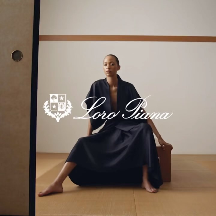 Elegant adult female model in a dark blue open-front dress, seated indoors against an off-white wall, with a logo visible.