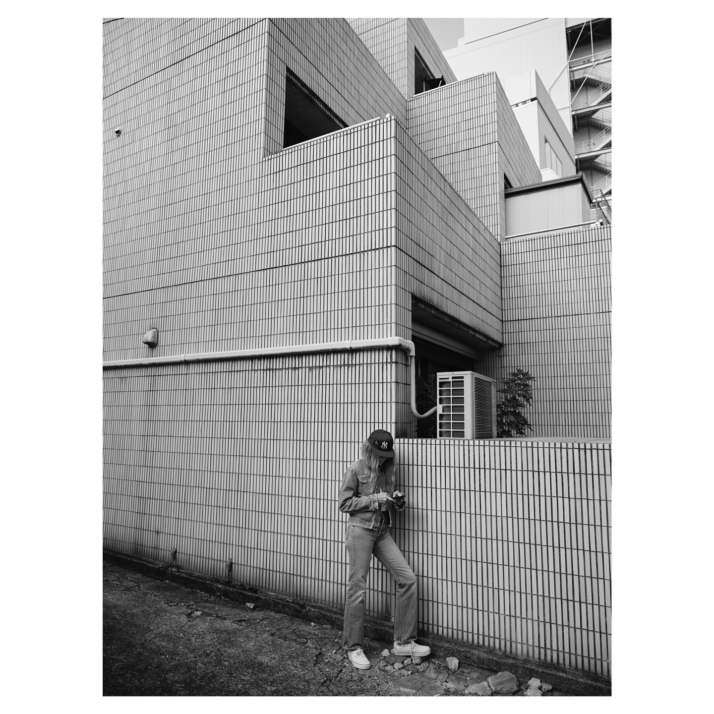 Monochrome full-length portrait of a person in a cap and denim standing against a tiled building exterior.