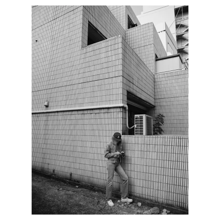 Monochrome full-length portrait of a person in a cap and denim standing against a tiled building exterior.