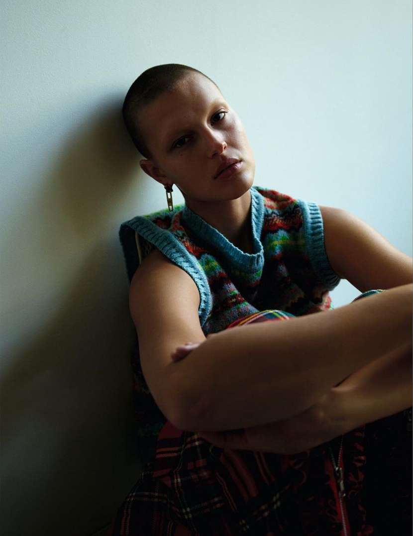 Indoor portrait of a shorthaired young adult female wearing a colorful knit vest. She is posed with arms crossed, leaning against a wall.