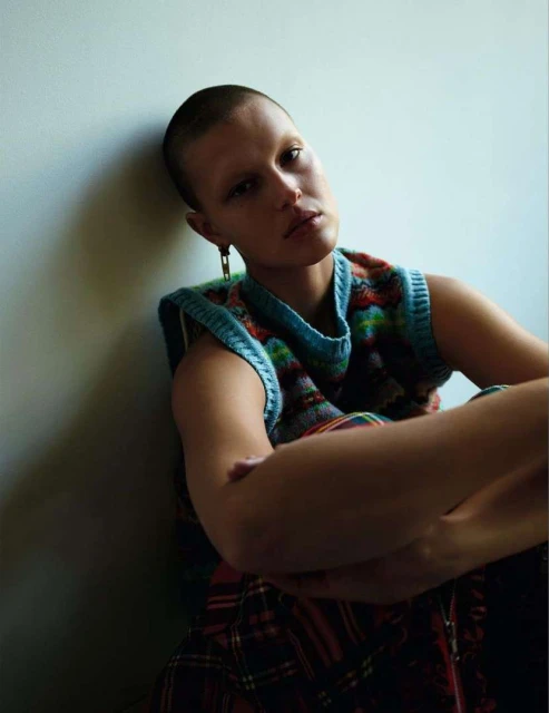 Indoor portrait of a shorthaired young adult female wearing a colorful knit vest. She is posed with arms crossed, leaning against a wall.