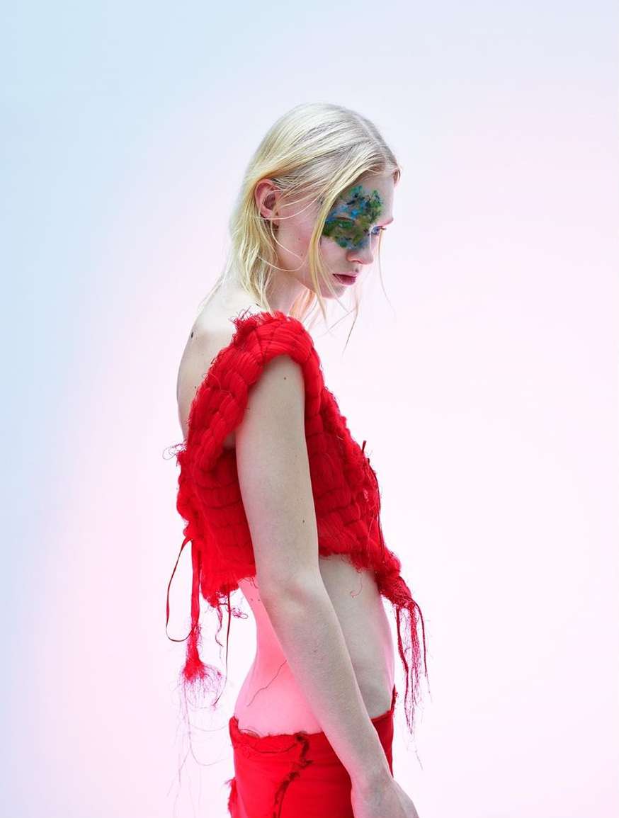 Blonde fashion model in red knit top and pants poses in a side profile against a light gradient background. Face paint detail.